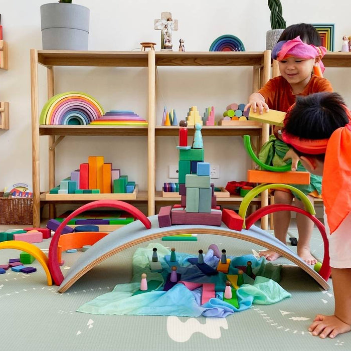 Two children playing with colourful wooden Grimm's rainbow arches and Sarah's Silks playsilks on a play mat in a bright children's room. Shelves in the background hold books, toys, and decor.