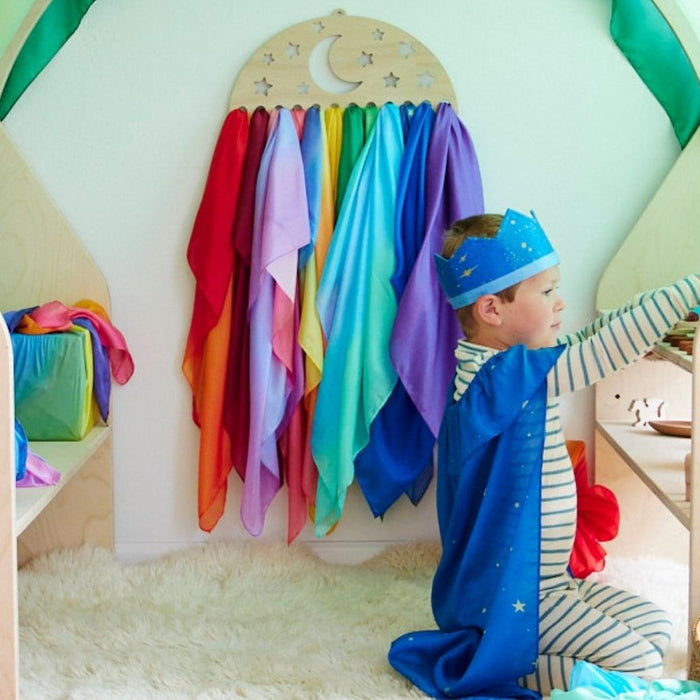 Child engaging in open-ended imaginative play in a wooden cubby draped with a giant Sarah's Silks playsilk and a selection of playsilks hanging up in the background