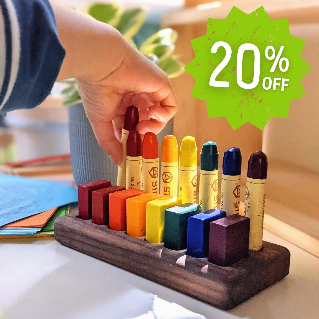 Colourful STOCKMAR crayons on a wooden stand with a hand reaching for one, promotional badge indicating 20% off.