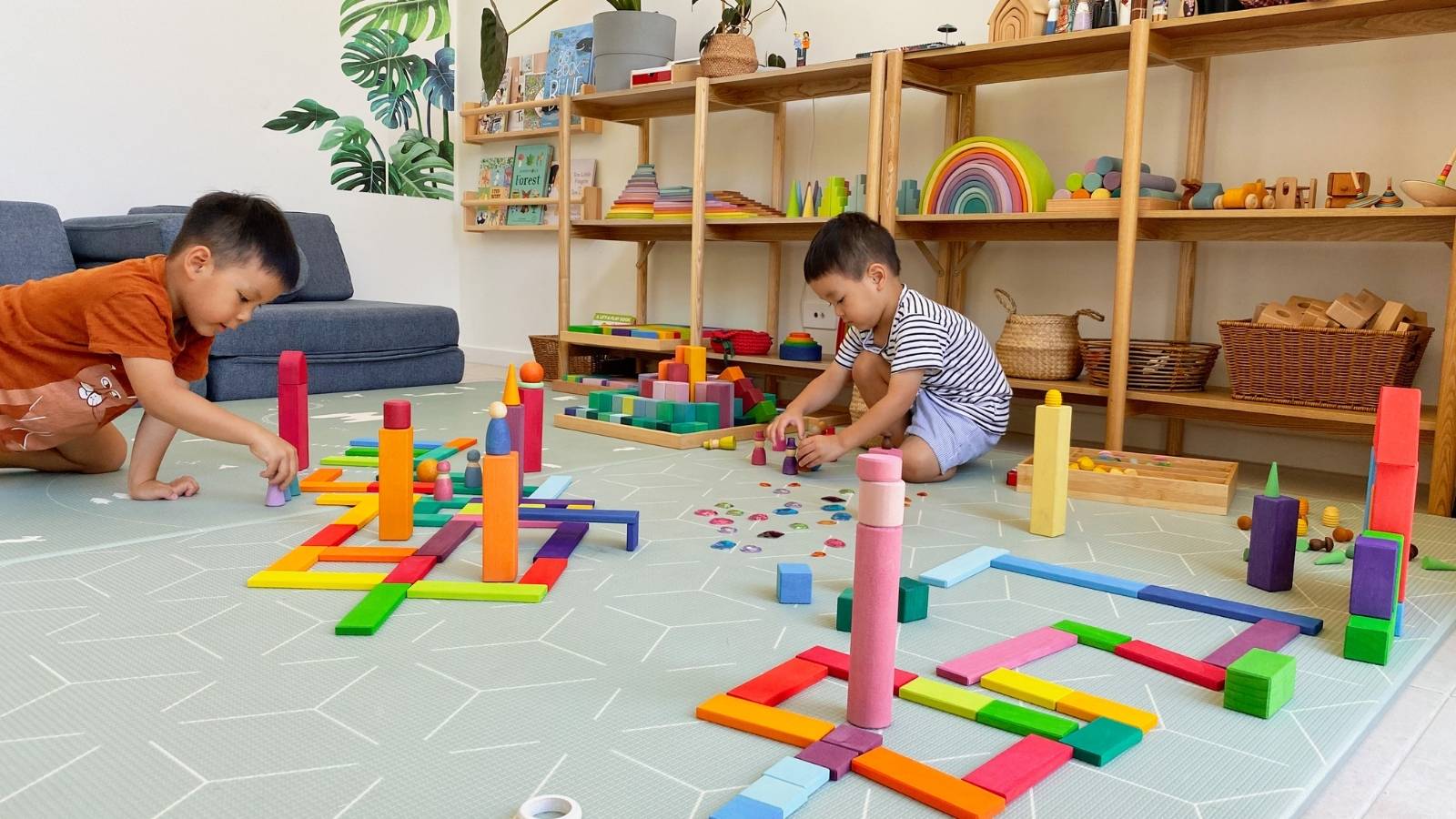 Two children playing with colourful building blocks on a play mat in a room with shelves of open-ended toys