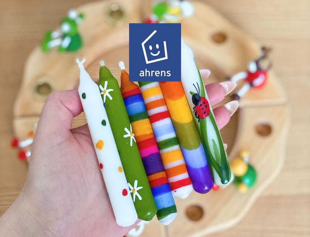 Ahrens candles lifestyle image with logo, featuring a set of colourful, hand-painted taper candles in front of a wooden celebration ring – Oskar's Wooden Ark