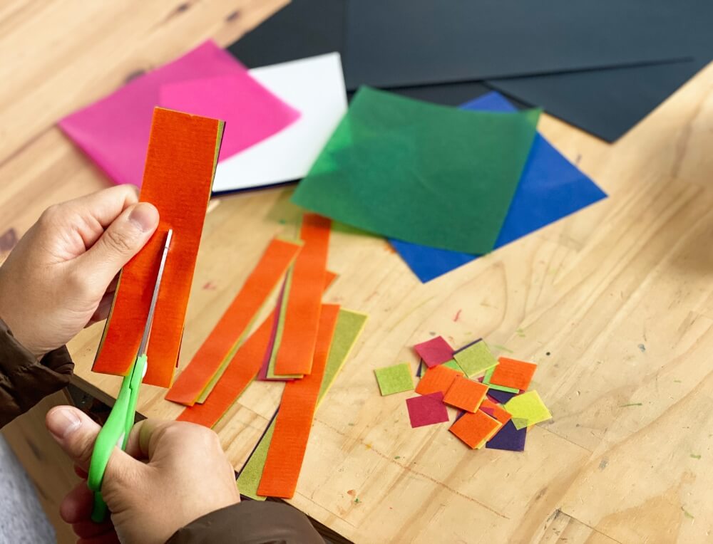 Materials for making rainbow sun catchers including coloured waxed kite paper, black cardstock, and clear contact