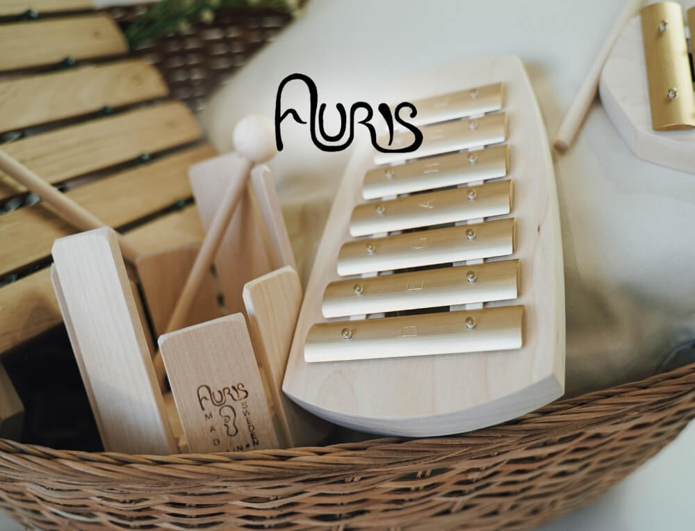 Auris Music lifestyle image with logo, featuring a wooden xylophone toy inside a wicker basket, with other wooden musical toys around it – Oskar's Wooden Ark