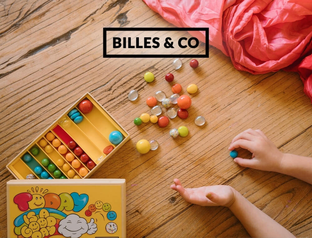 Billes & Co Marbles lifestyle image with logo, featuring scattered glass marbles, and a child's hands playing – Oskar's Wooden Ark