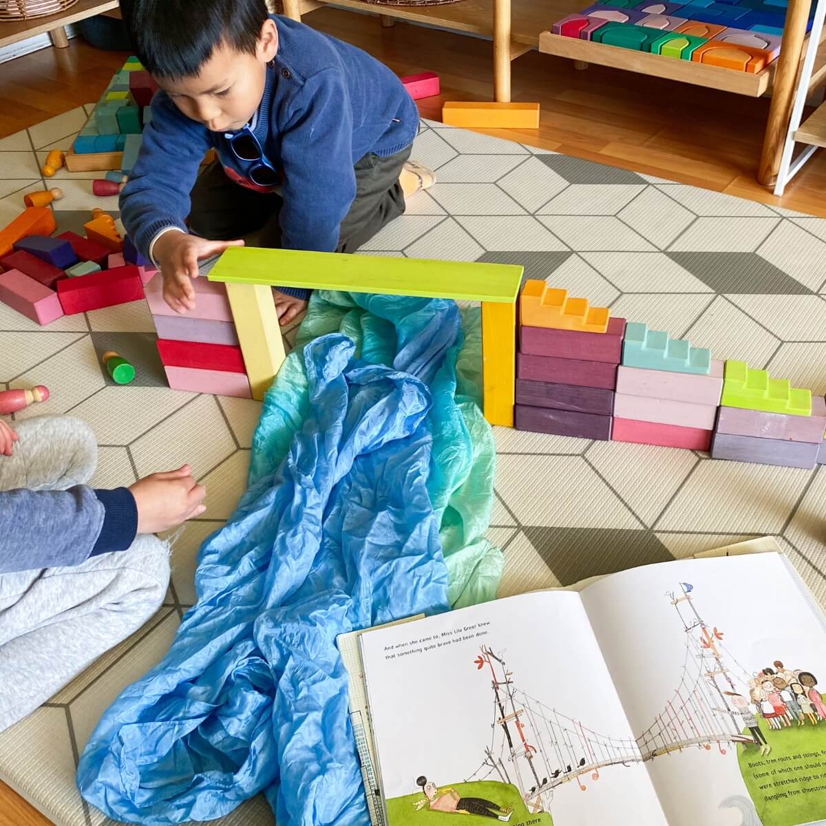 A young child kneels on a patterned play rug in a preschool or daycare setting, interacting with colourful wooden blocks and a bright blue and green play silk - building a bridge, exploring a scene from a children's book through play