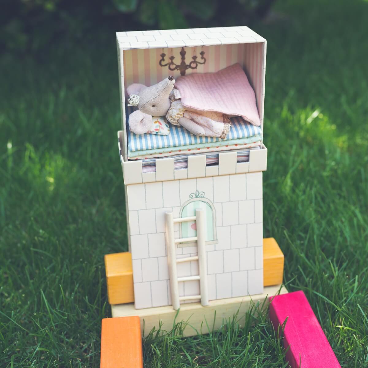 A miniature Maileg dollhouse tower with a small princess mouse doll in a bed on top, set outdoors on green grass with coloruful wooden blocks - The Princess and the Pea bookish play setup.