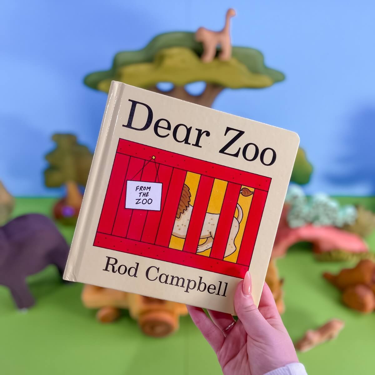 A hand holds a children's board book titled 'Dear Zoo' by Rod Campbell. In the background is a bookish play setup with wooden toy animal figures and a tractor toy from Oskar's Wooden Ark.