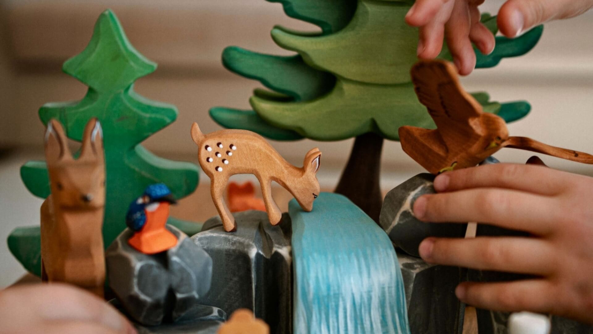 Children's toy forest scene with Bumbu wooden animals and trees, featuring a child's hands interacting with the toys.
