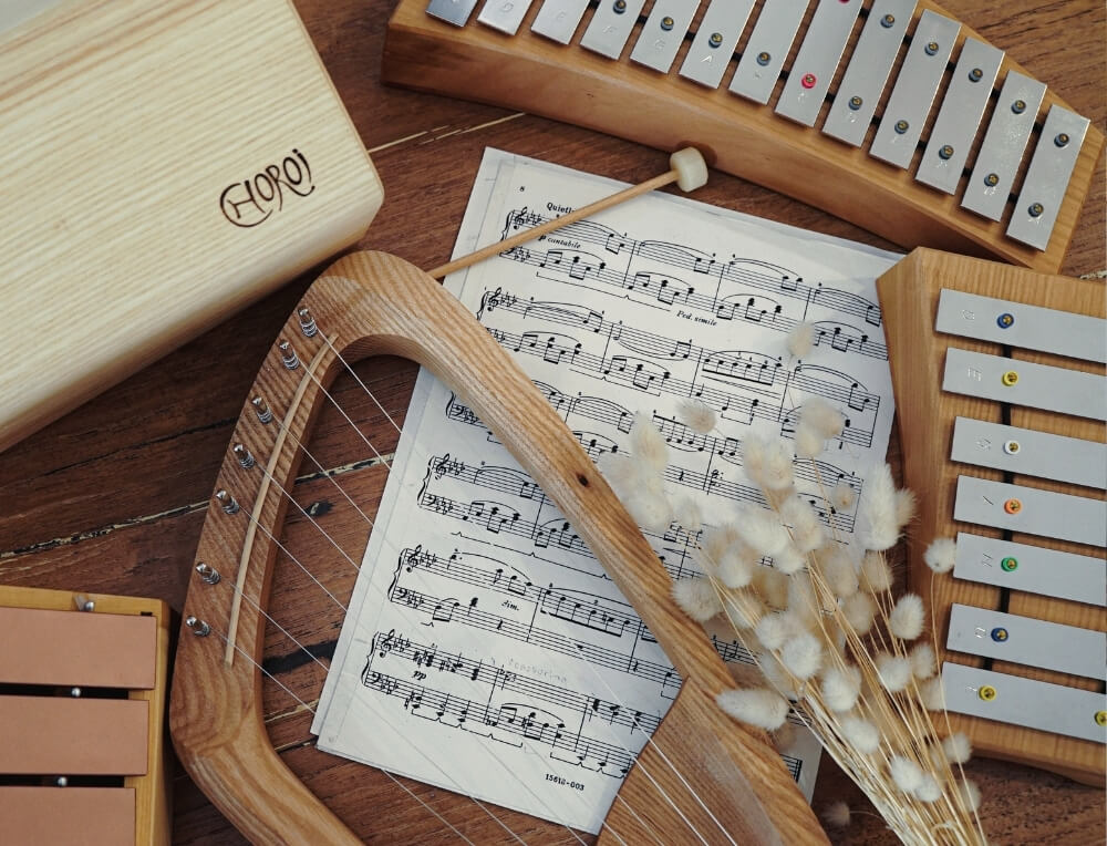 Choroi Music lifestyle image with logo, featuring a wooden xylophone , metallophone and a wooden harp arranged on a wooden surface with printed sheet music  – Oskar's Wooden Ark
