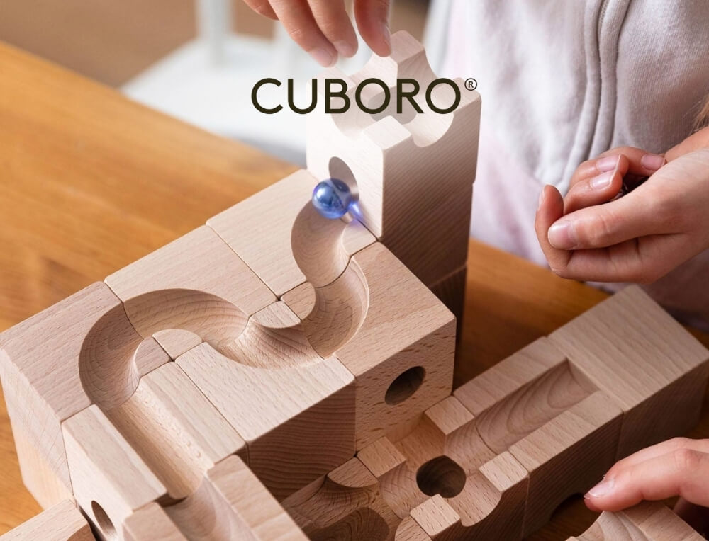 CUBORO lifestyle image with logo, featuring a close-up of a person's hands assembling a CUBORO wooden marble run with interconnecting blocks on a wooden table. A blue marble is sliding down a carved track – Oskar's Wooden Ark