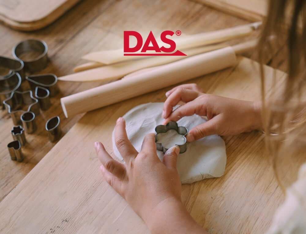 DAS Clay lifestyle image with logo, featuring a child's hands using a metal flower-shaped clay cutter on white clay over a wooden table. Several other metal cookie cutters are lined up on the left, with wooden rolling pins in the background – Oskar's Wooden Ark