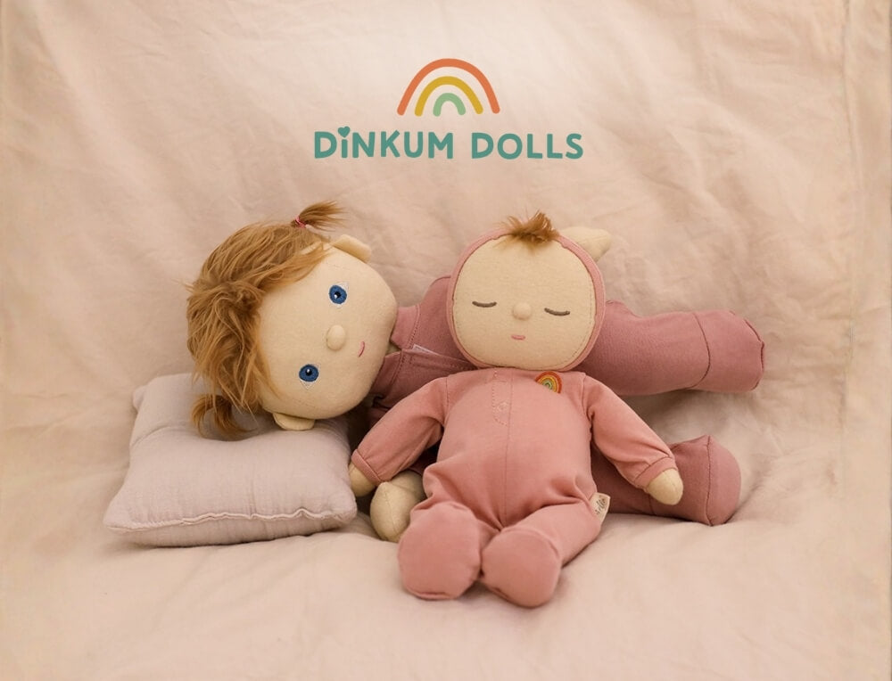 Dinkum Dolls by Olli Ella lifestyle image with logo, featuring two Dinkum Dolls plush dolls posed on a soft beige fabric backdrop with a pillow. One doll lies closed-eyed in a pink outfit, and the other reclines with blonde hair and blue eyes wearing a light pink top – Oskar's Wooden Ark