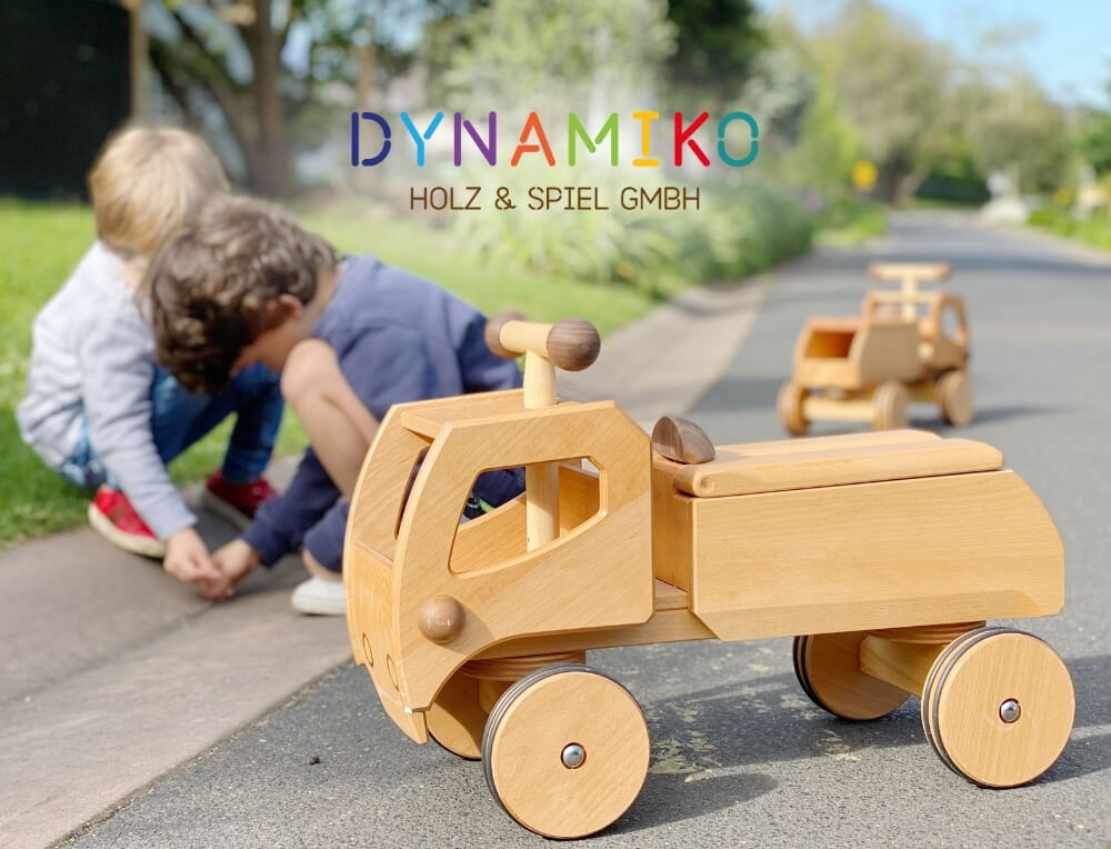 Dynamiko lifestyle image with logo, featuring two children playing on a street corner with wooden ride-on car toys – Oskar's Wooden Ark