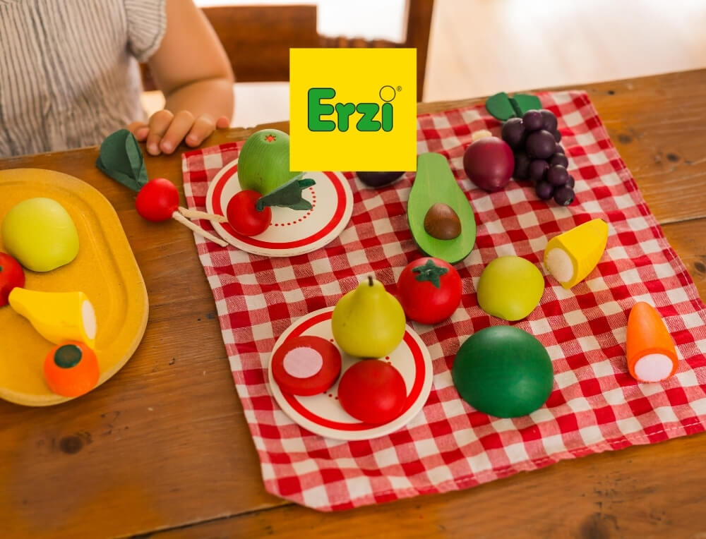 Erzi lifestyle image with logo, featuring a children's wooden toy fruit and vegetable play set arranged on a red-and-white checkered picnic blanket on a wooden table – Oskar's Wooden Ark