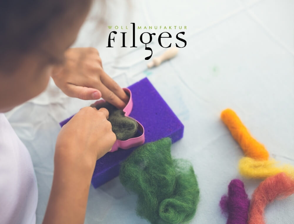 Filges lifestyle image with logo, featuring a child's hands felting a green butterfly shape with a pink metal cutter over a purple foam block, with colourful wool felt bundles nearby – Oskar's Wooden Ark