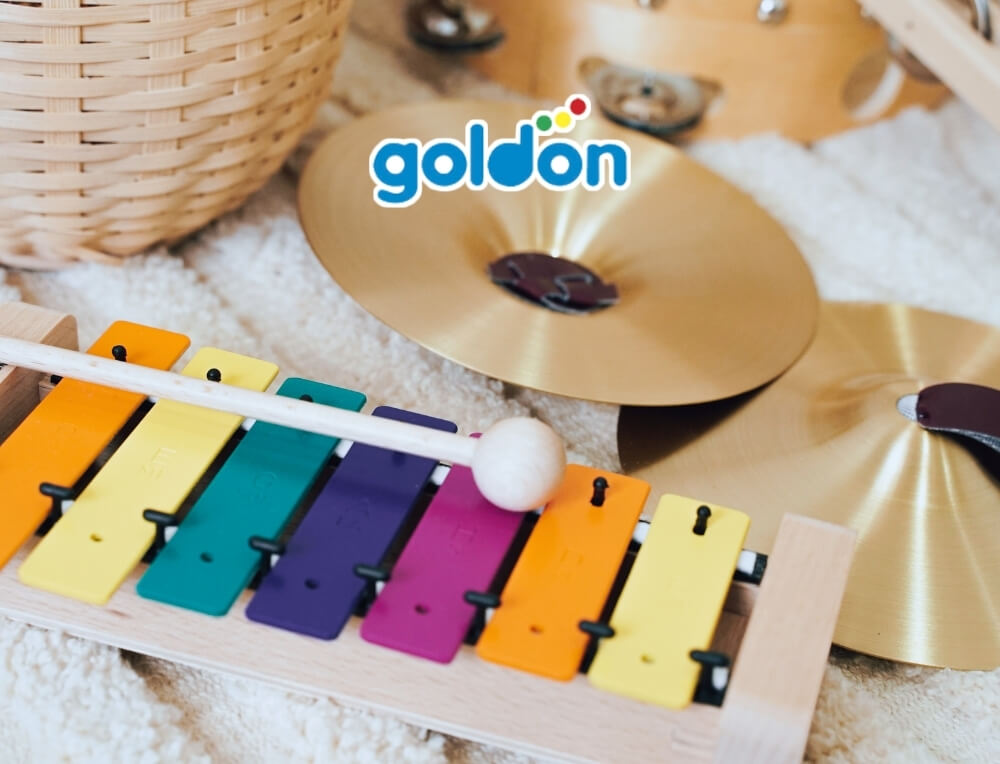 Goldon Music lifestyle image with logo, featuring a colourful wooden toy xylophone with rainbow-coloured keys and a wooden mallet, shown with gold cymbals on a soft white blanket – Oskar's Wooden Ark