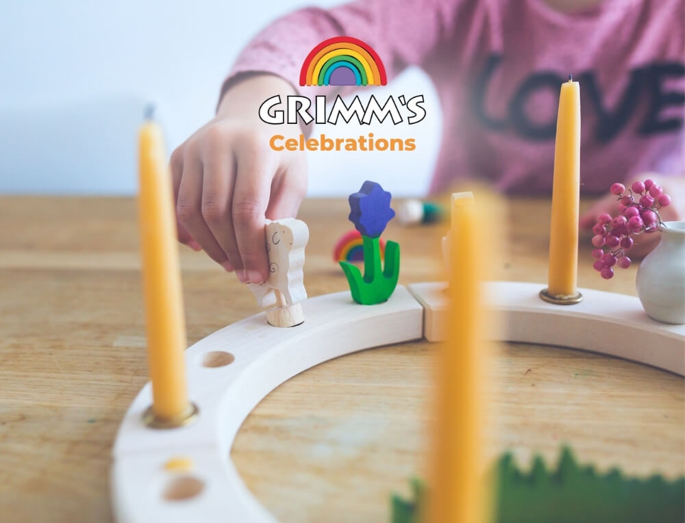 Grimm's Celebrations lifestyle image with logo, featuring a close-up of a child's hand placing a small wooden figurine into a celebration ring with yellow candles, on a wooden table – Oskar's Wooden Ark
