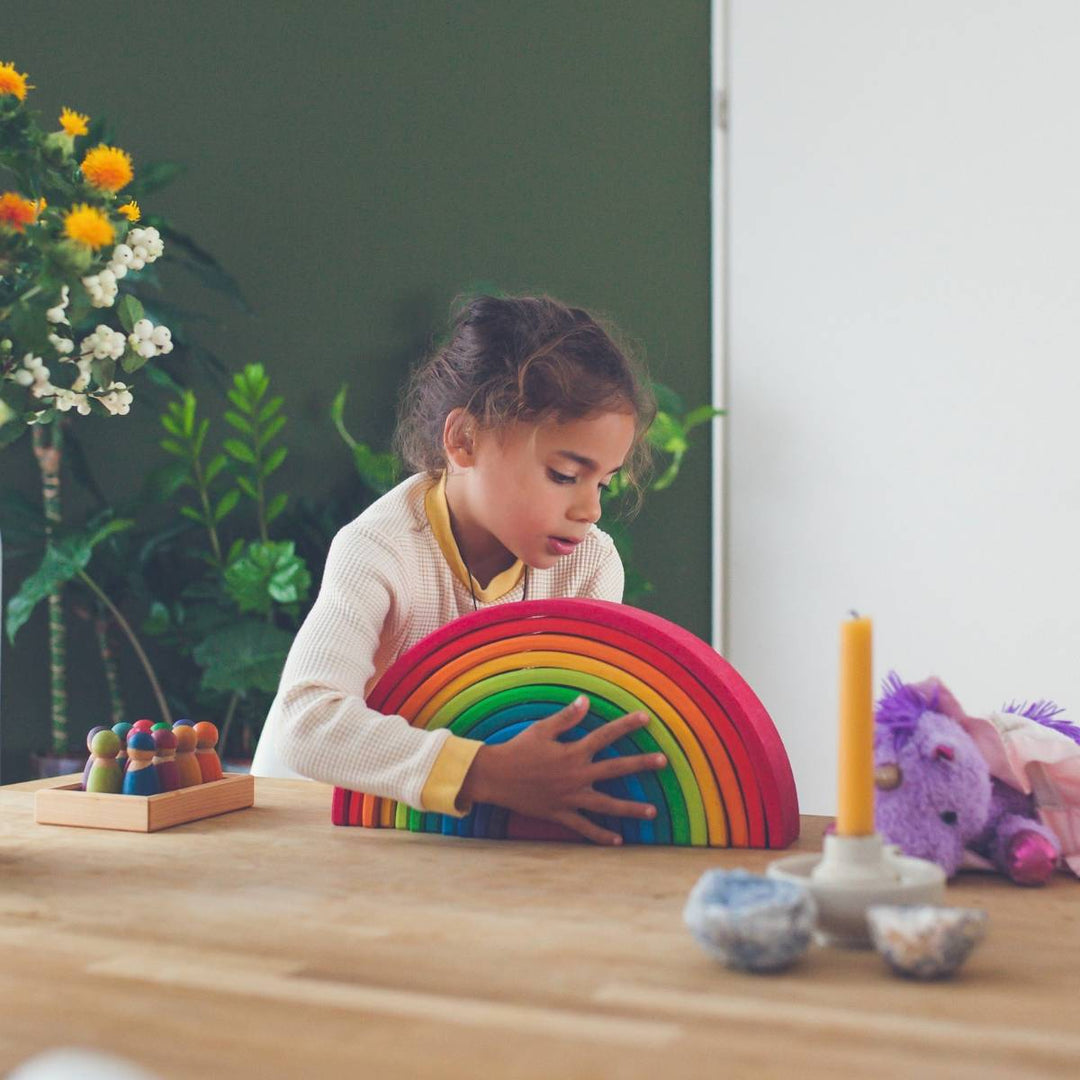 Child placing a Grimm's wooden rainbow on a table top alongside a set of Grimm's Rainbow Friends wooden peg dolls, ready to play