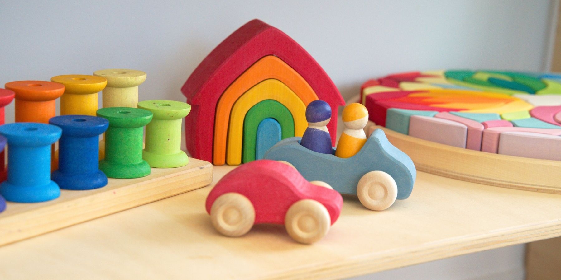 Colourful Grimm's wooden toys including cars, a house, and spools on a light playroom shelf surface.