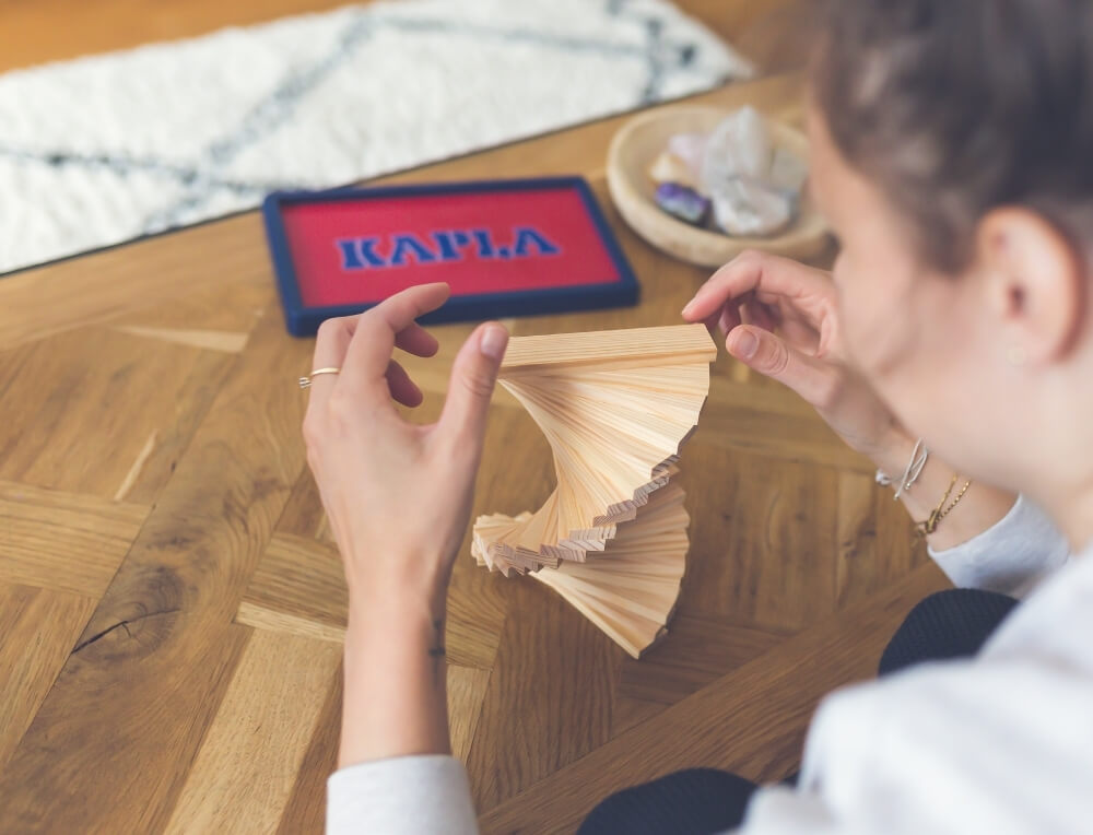KAPLA lifestyle image with logo, featuring a person building a spiral structure with KAPLA construction planks over a wooden table – Oskar's Wooden Ark