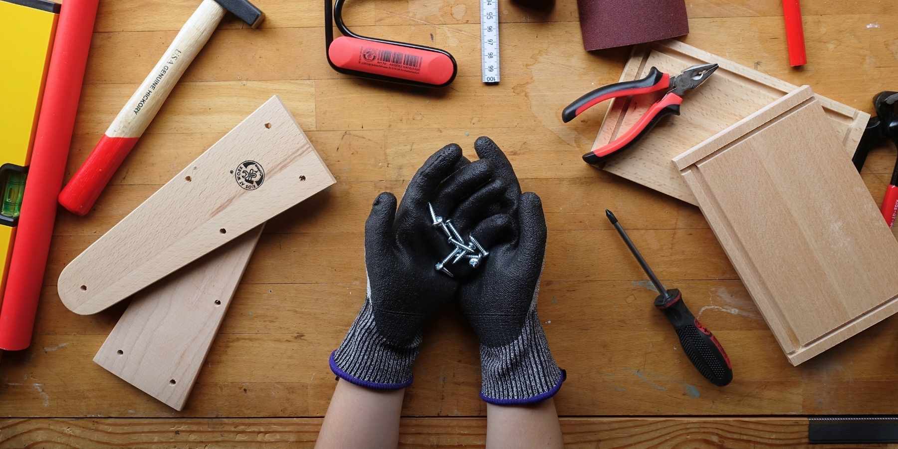 Pair of children's hands wearing black gloves holding screws and nails on a wooden surface with Kids at Work children's woodworking tools and materials.