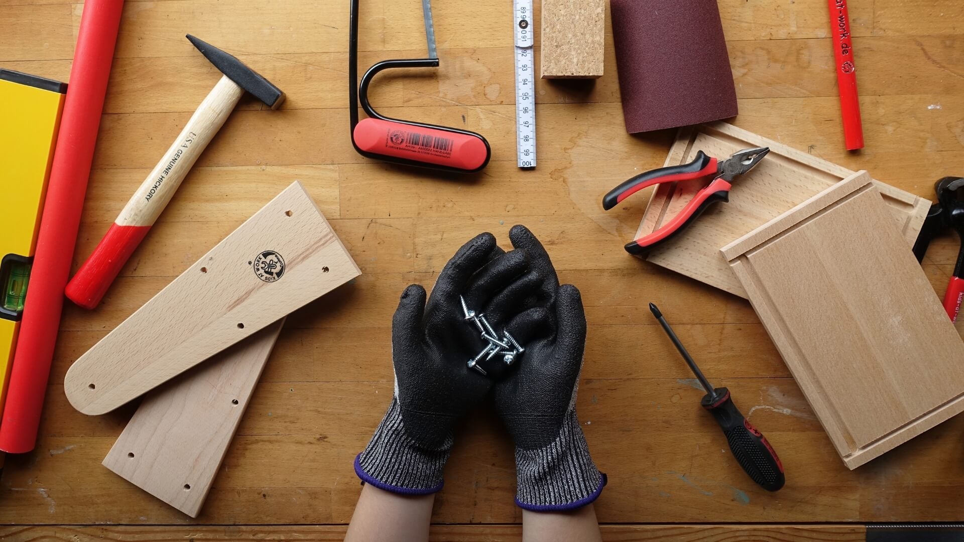 Child wearing wood working gloves surrounded by various Kids at Work tools on a wooden workbench surface