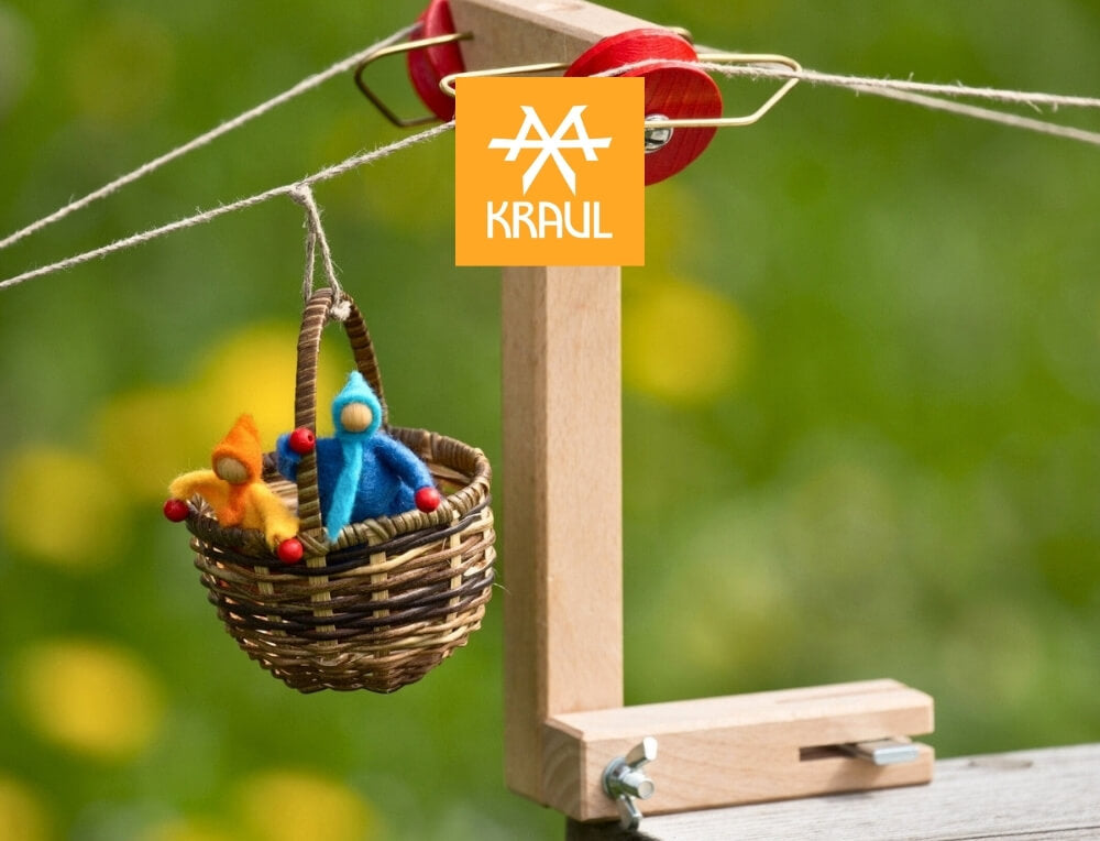 Kraul STEM  lifestyle image with logo, featuring a small woven basket hanging from a wooden cable car frame on a rope, containing two tiny felt figures in blue and orange – Oskar's Wooden Ark