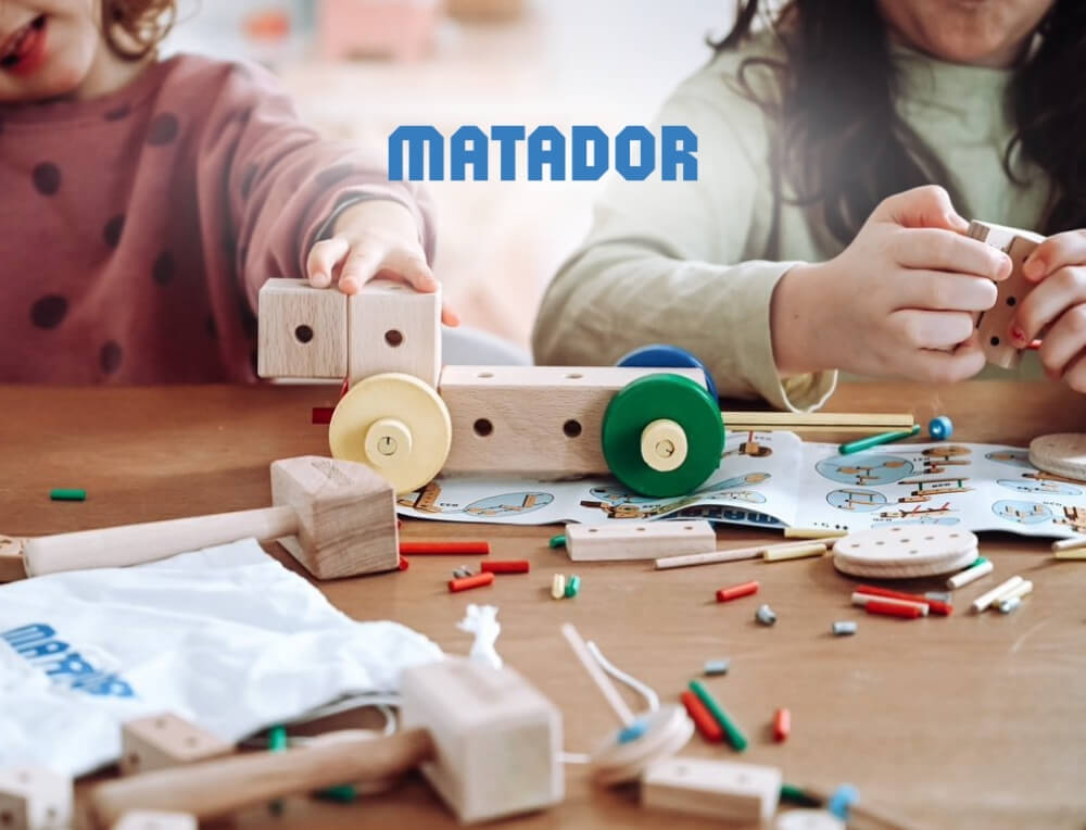 MATADOR lifestyle image with logo, featuring two children playing with a wooden toy construction set with colourful wheels and small wooden pieces - Oskar's Wooden Ark