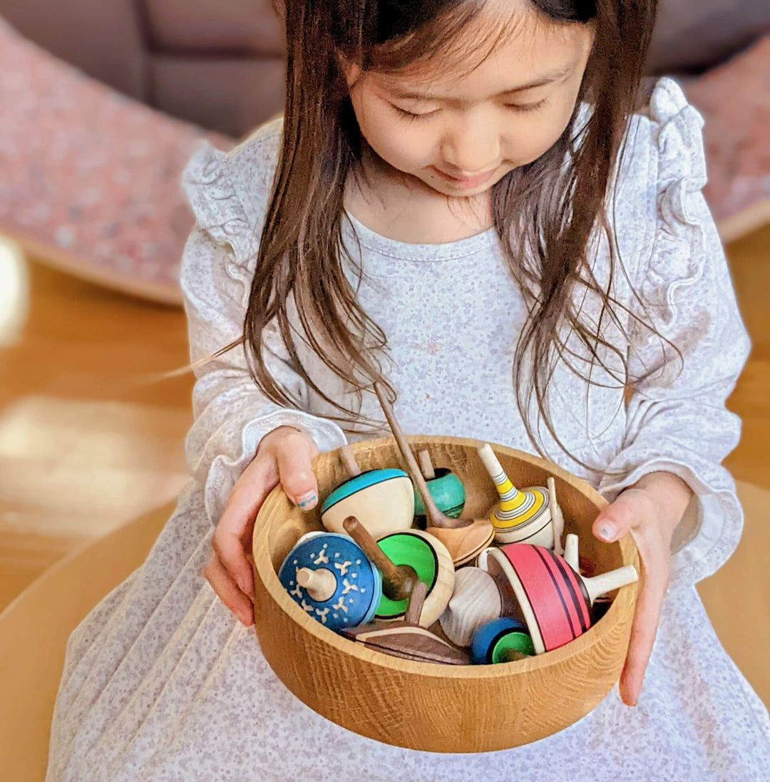 A young girl with long brown hair, wearing a light gray long-sleeve dress, holds a round wooden bowl filled with colorful wooden Mader spinning tops.