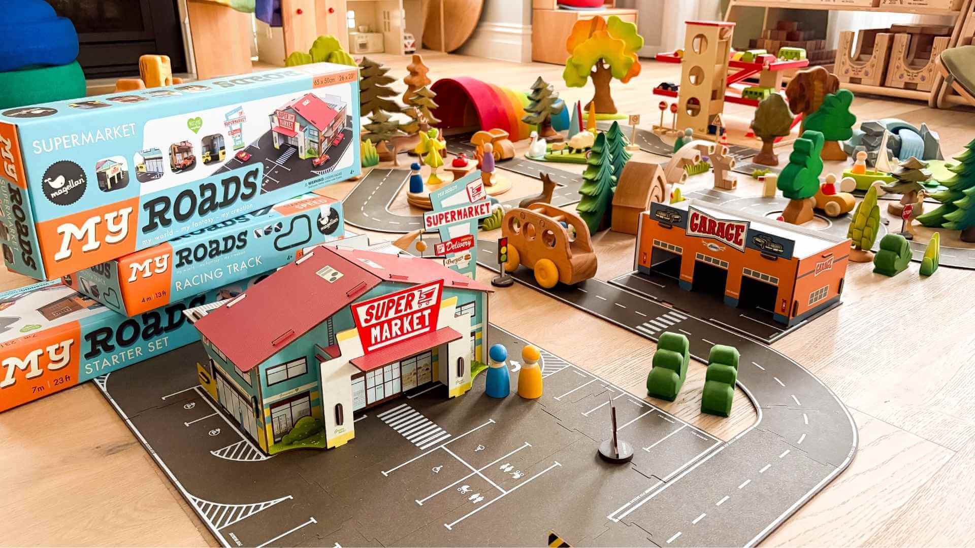 Toy town setup with buildings, cars, and 'Magellan My Roads' packaging on a wooden floor.