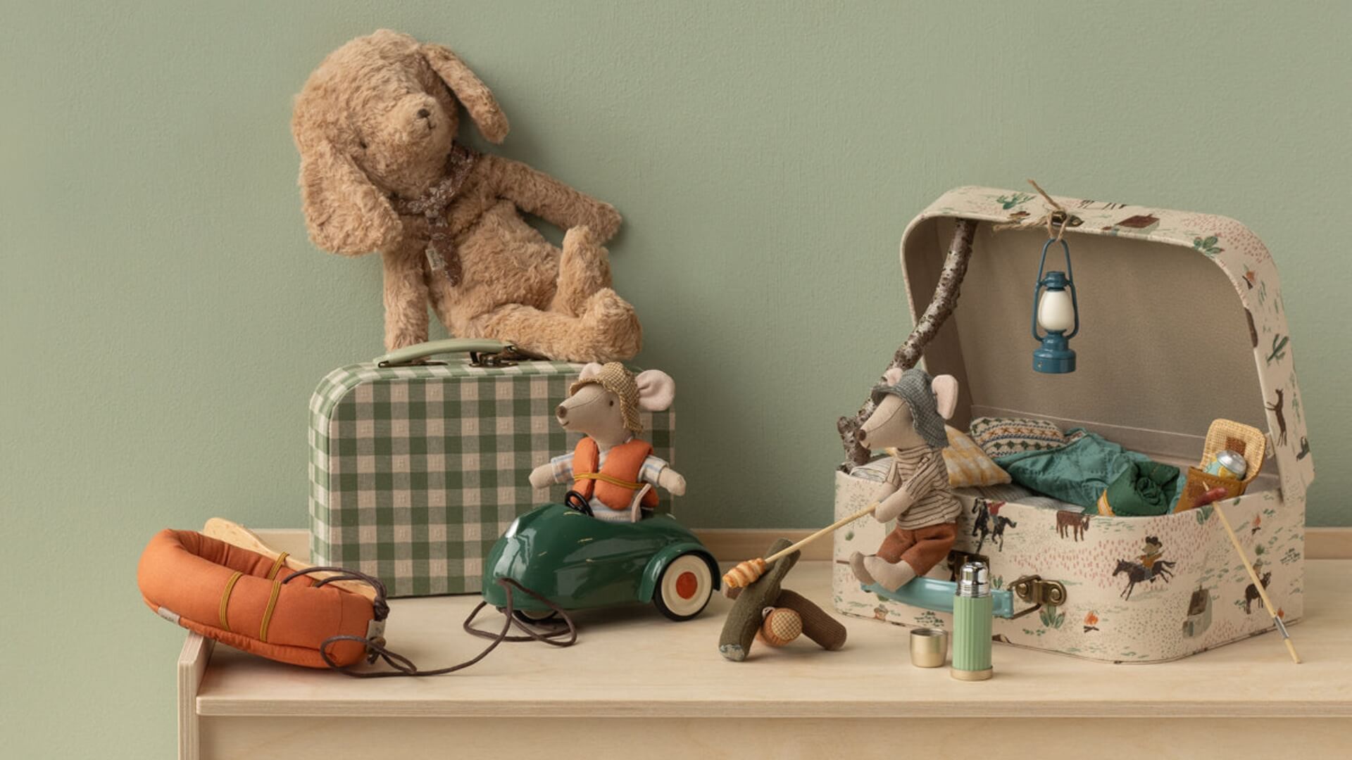 Collection of Maileg toys including a teddy bear, mouse on a car, and a suitcase on a shelf with a green wall background.