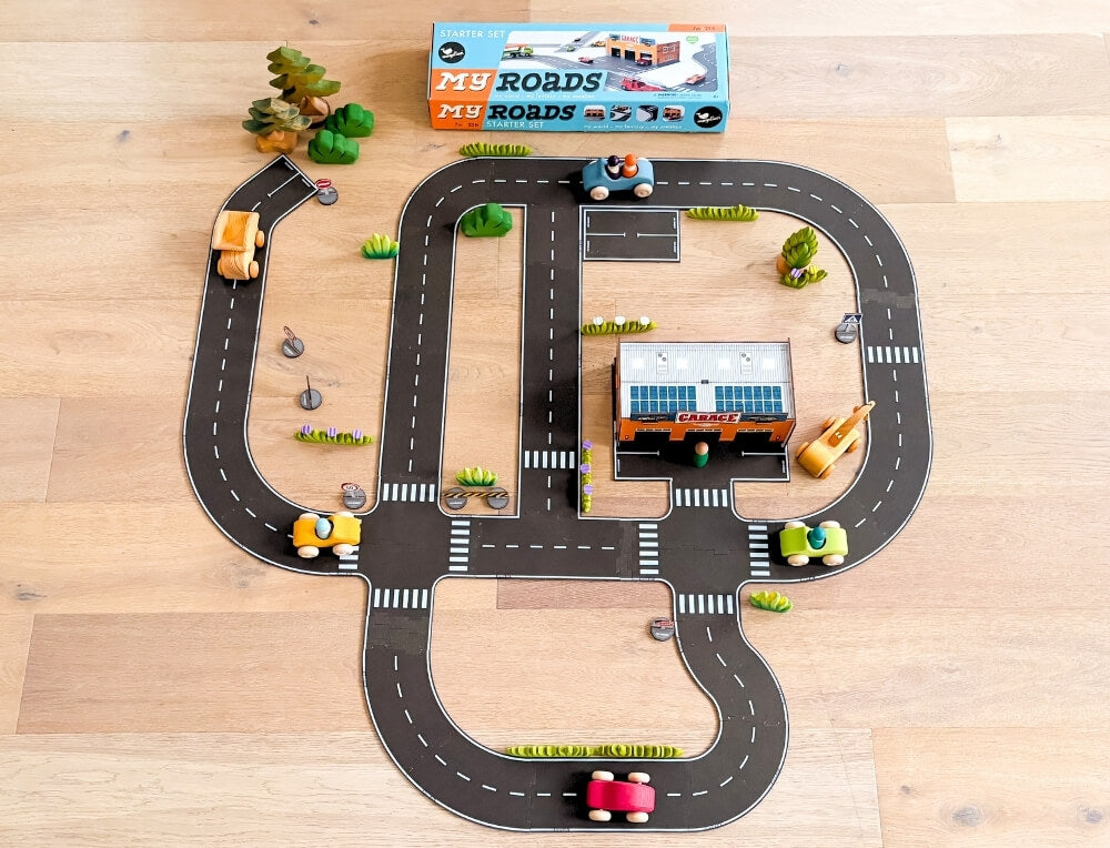 My Roads lifestyle image with logo, featuring a toy road playset with curved black roads, white dashed lines, small green trees, miniature buildings, and colourful toy cars – Oskar's Wooden Ark