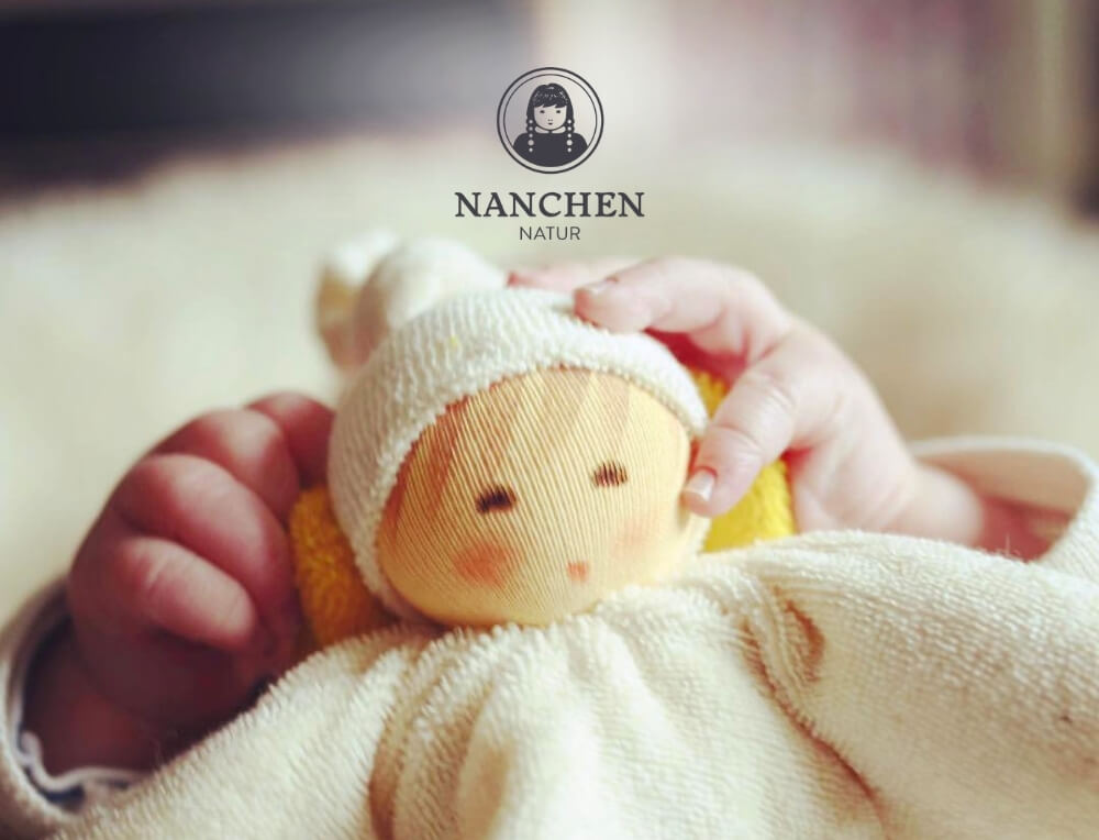 Nanchen Natur lifestyle image with logo, featuring a close-up of a baby's hands holding a small knitted doll – Oskar's Wooden Ark