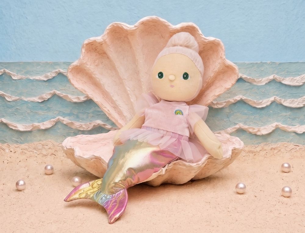 A mermaid Dinkum Doll with a pink dress and iridescent tail sits inside a large pink seashell on a sandy beach set with blue ocean waves and pearls - Oskar's Wooden Ark