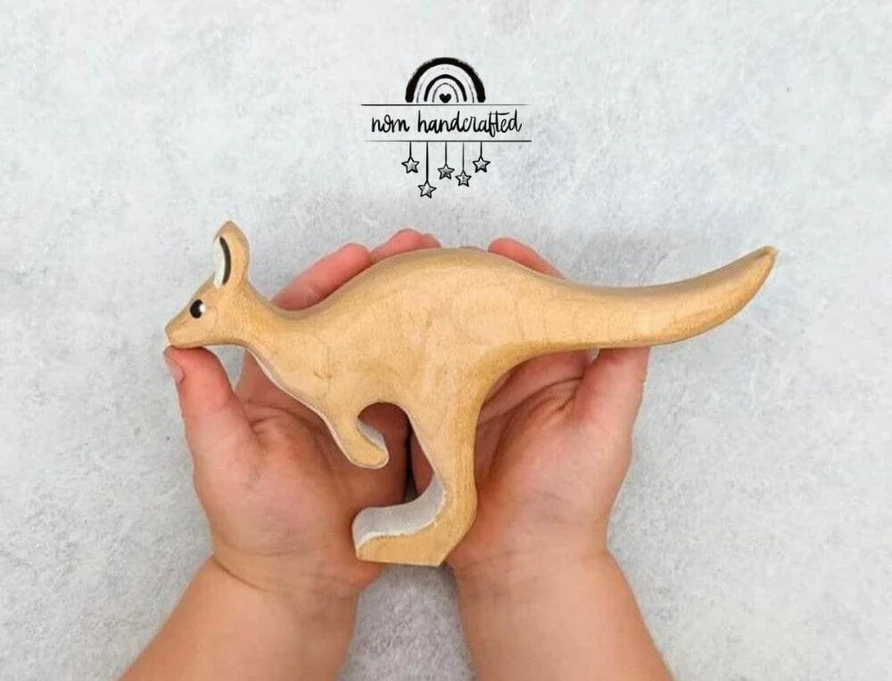 NOM Handcrafted lifestyle image with logo, featuring a wooden kangaroo toy being held in a child's hands – Oskar's Wooden Ark
