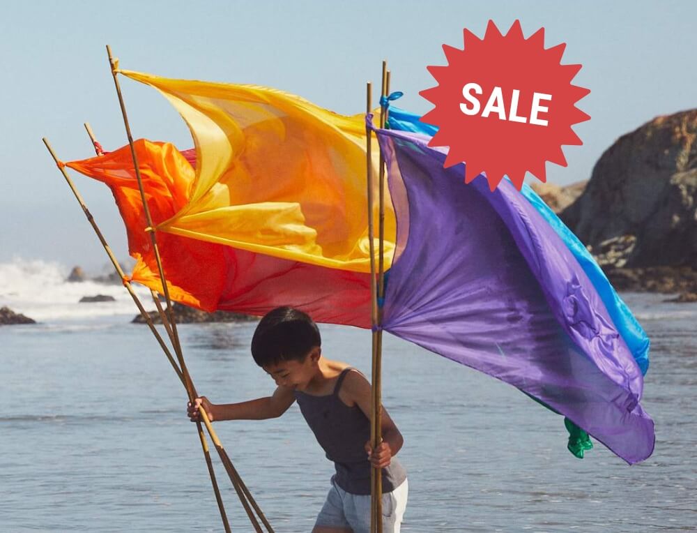 Image of a child holding Sarah's Silks flags on the beach with the word 'SALE' - Wooden toys and children's products on sale at Oskar's Wooden Ark