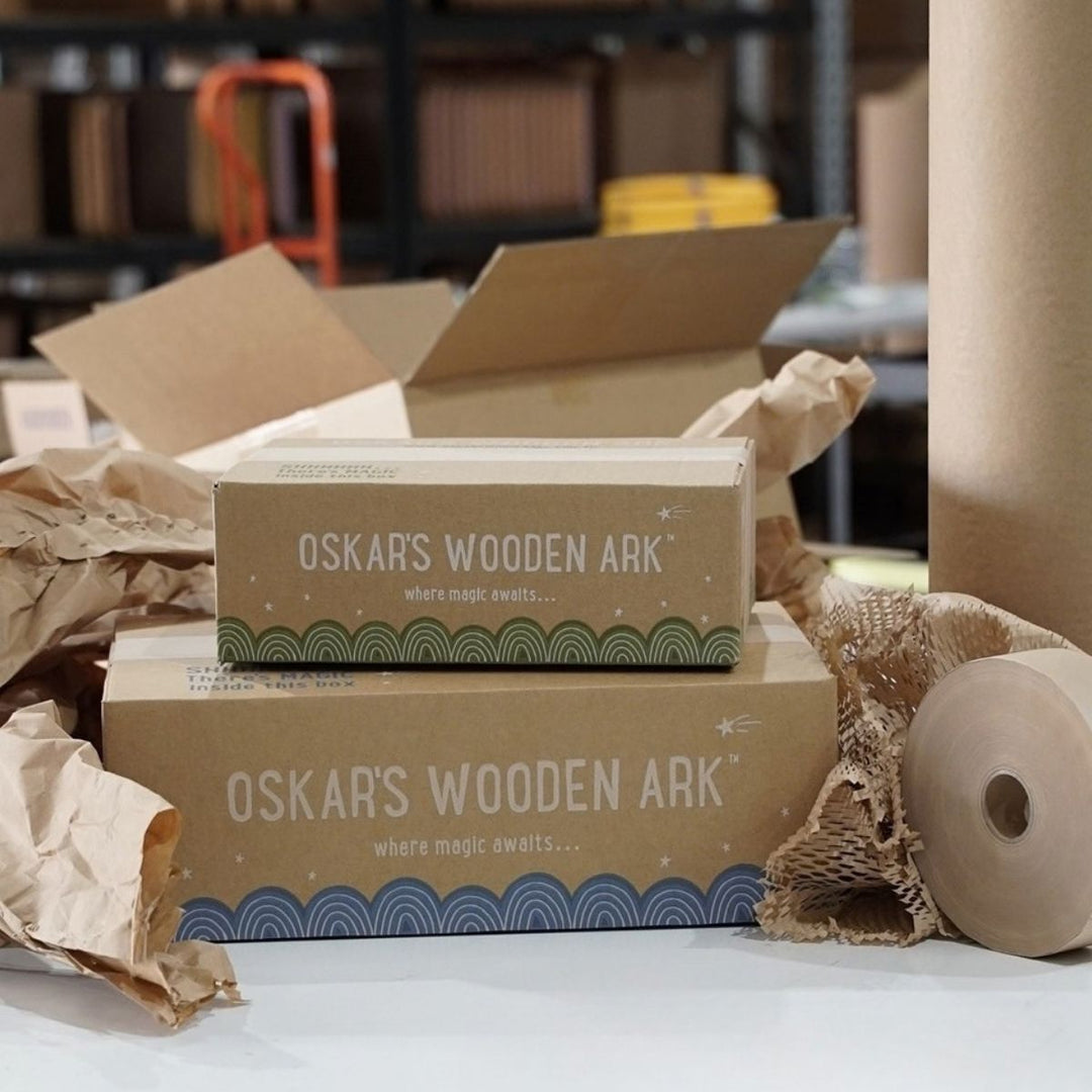 Two brown kraft paper boxes branded 'OSKAR'S WOODEN ARK' are stacked on a table with crumpled brown paper and burlap around them, with an open cardboard box and warehouse shelves blurred in the background.