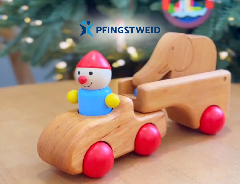 Pfingstweid Wooden Toys lifestyle image with logo, featuring a wooden toy car with a small painted clown figure in the driver's seat and a wooden elephant in a trailer – Oskar's Wooden Ark