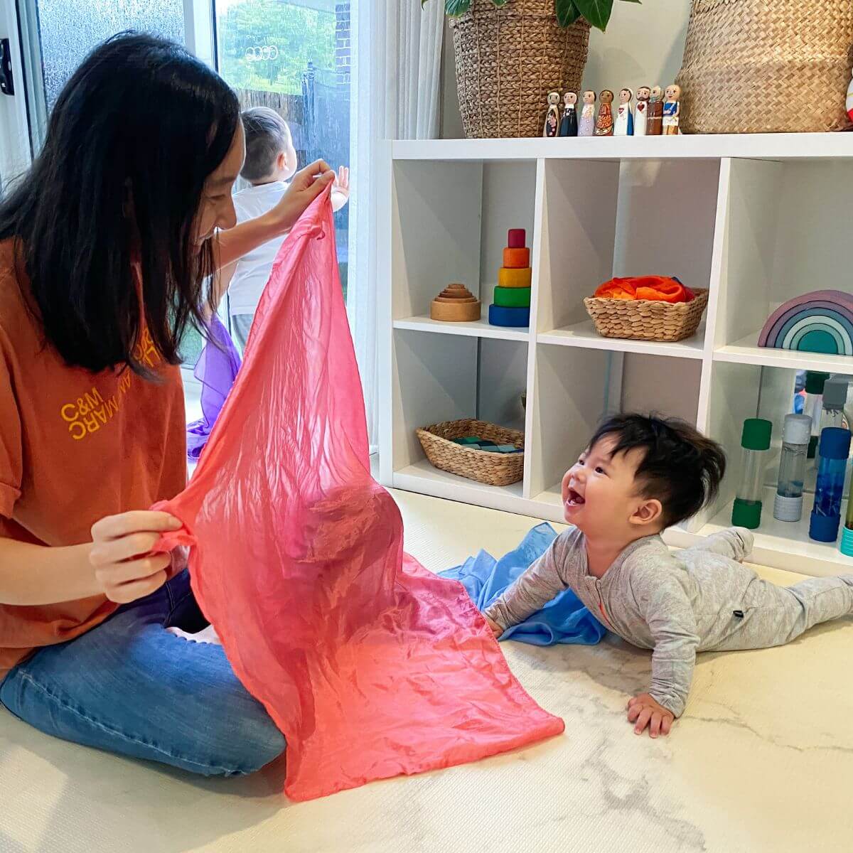 A woman and a baby play peekaboo with a large pink Sarah's Silks Playsilk in a bright indoor room with white shelves and toys.