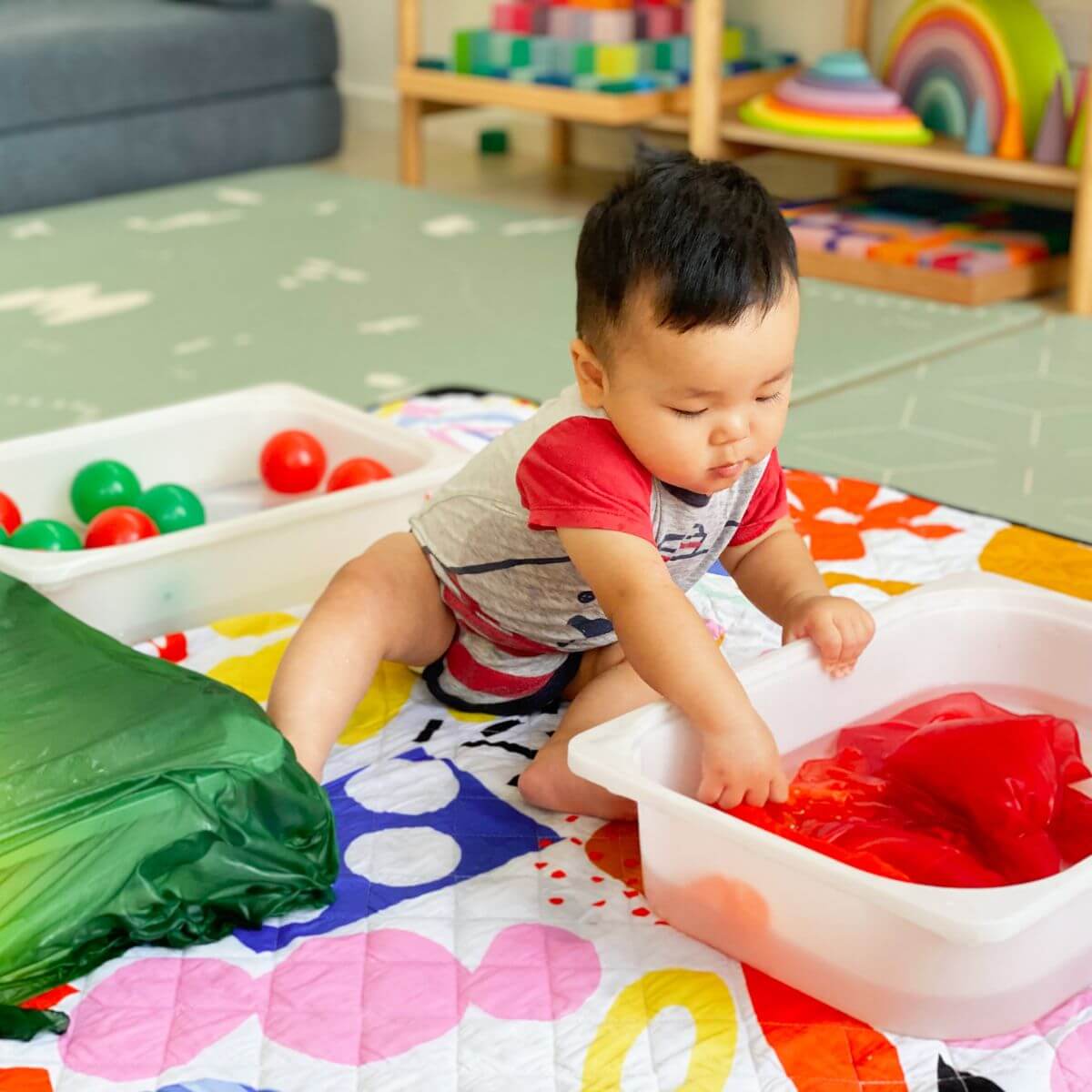 A baby sitting on a colorful play mat, reaching into a white plastic tub filled with water and a red Sarah's Silks Playsilk, with toy bins and shelves in the background.