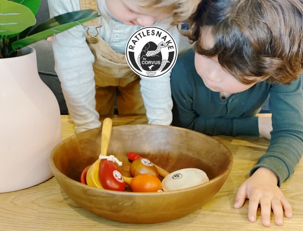 Rattlesnake by Corvus lifestyle image with logo, featuring two children leaning over a wooden bowl filled with percussion instruments – Oskar's Wooden Ark