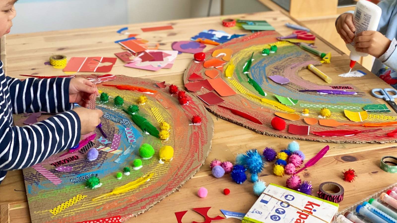 Children crafting colourful rainbows using Kitpas crayons, recycled cardboard and collage materials. One child is visible on the left and another is visible on the right, working on the projects.