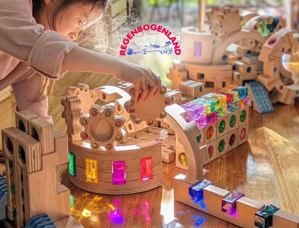 Regenbogenland lifestyle image with logo, featuring a child building a tower with rainbow coloured translucent gems and wooden building blocks – Oskar's Wooden Ark
