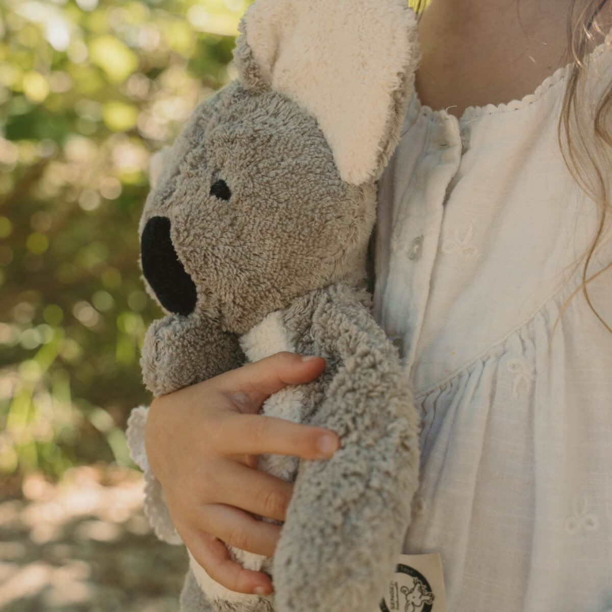 A child in a white dress holds a limited edition SENGER koala toy outdoors with a soft, blurred green and yellow background  - Oskar's Wooden Ark Australia