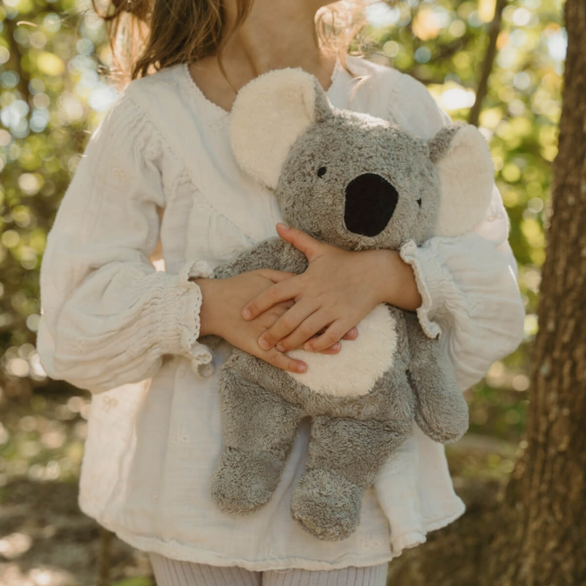 A child in a white blouse holds a plush limited edition SENGER  koala toy outdoors in a sunlit Australian forest setting.