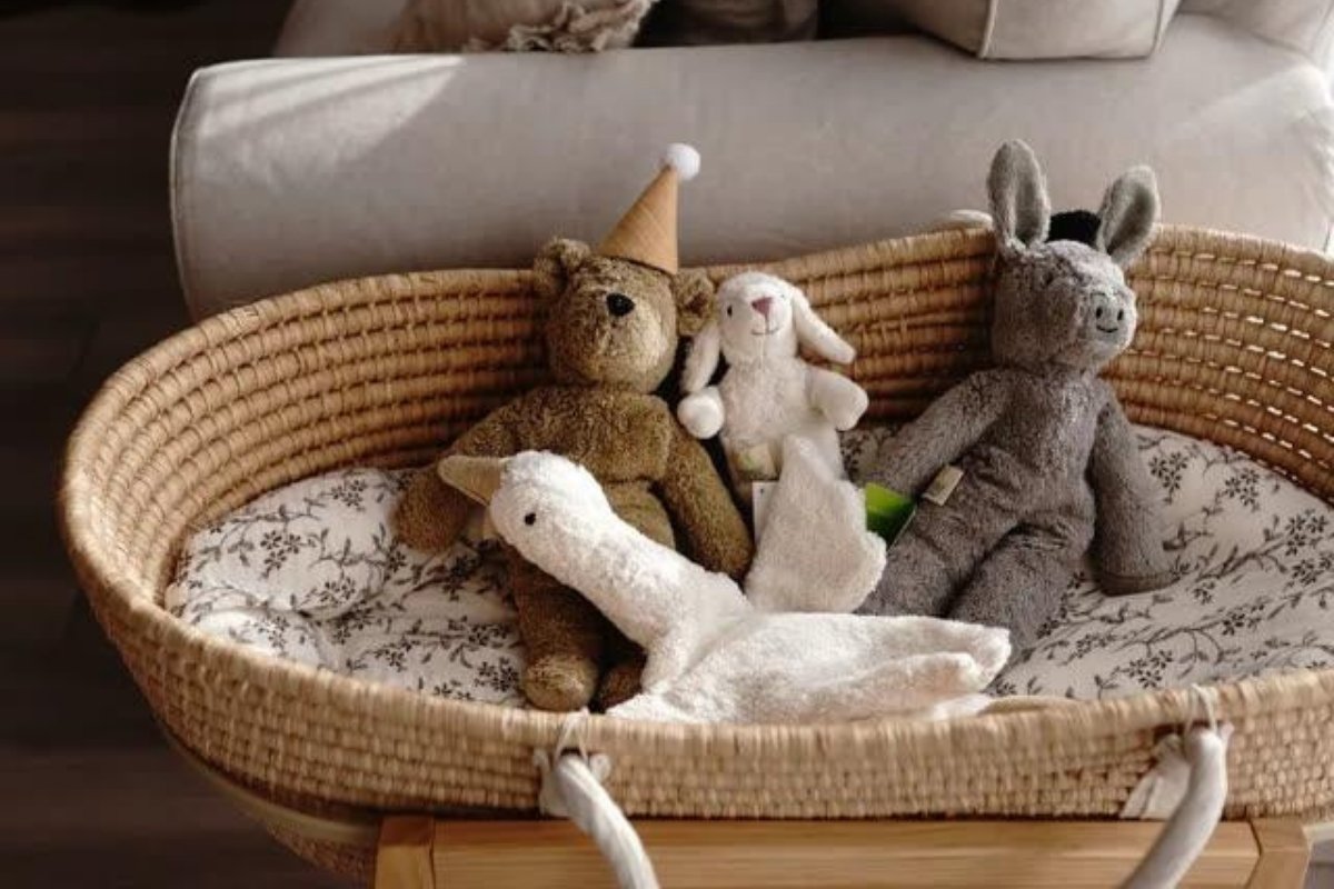 A woven natural baby bassinet with a floral-patterned cushion, holding four SENGER stuffed toys including a teddy bear, lamb, goose, and donkey.