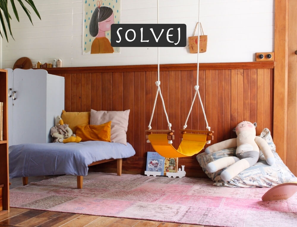 Solvej swings lifestyle image with logo, featuring a cosy children's room corner with a wooden wall, pink rug, hanging yellow swing chair – Oskar's Wooden Ark