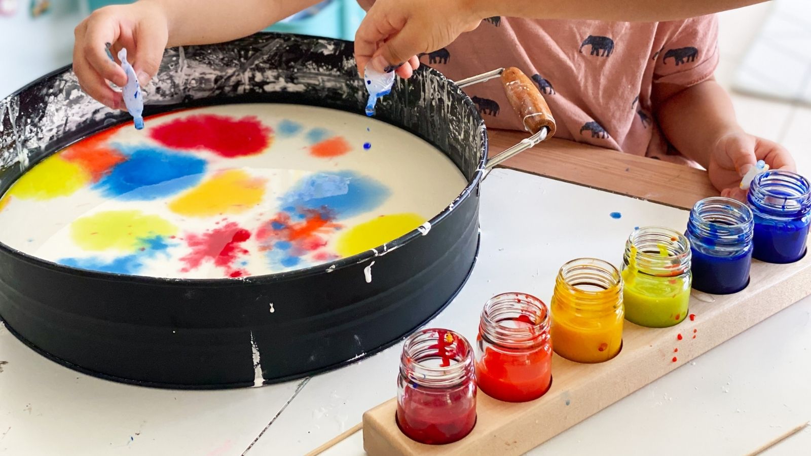 A child's hands and arms are visible as they participate in a colorful art activity, dripping blue paint into a large black tub filled with oobleck and splashes of red, yellow, and blue. A wooden tray holds several small glass jars containing red, orange, yellow, green, blue, and purple STOCKMAR watercolour paints on a white table.