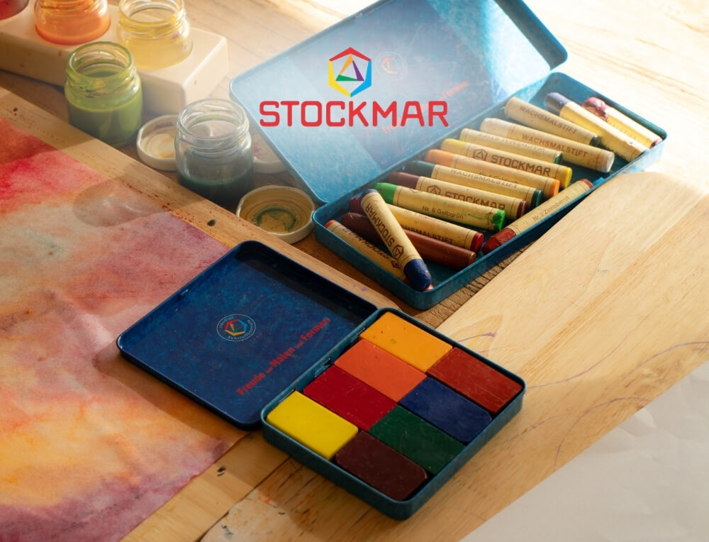 STOCKMAR Art lifestyle image with logo, featuring blue metal tins with block and stick crayons alongside small paint jars and a watercolour sheet – Oskar's Wooden Ark