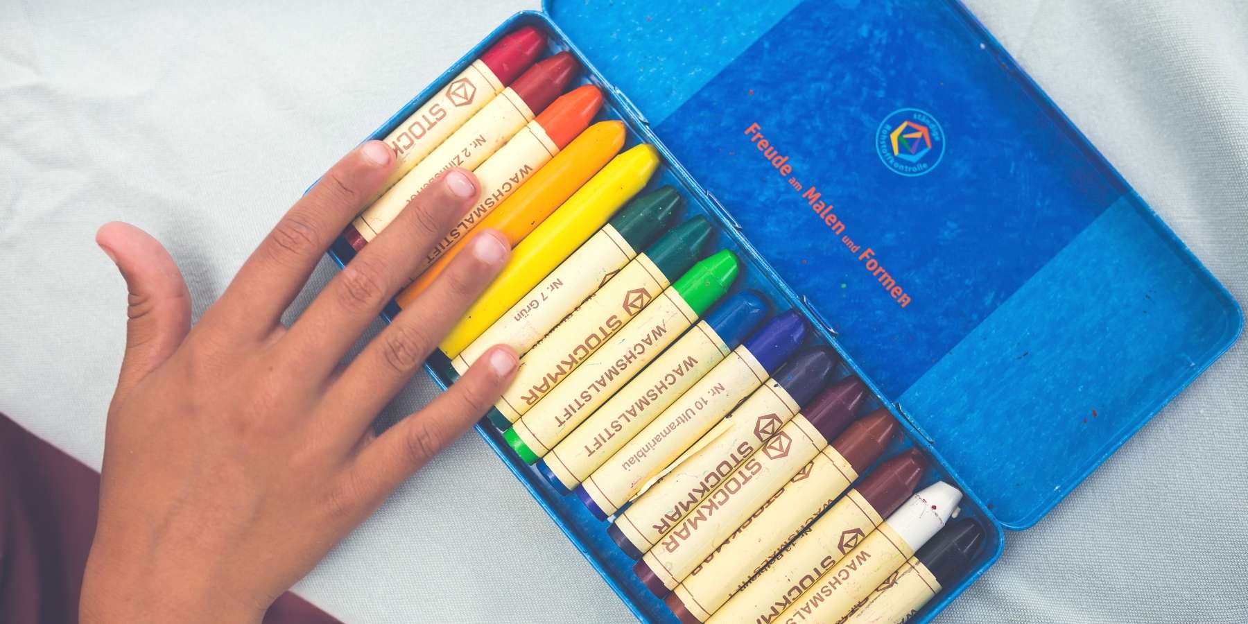 Box of colourful STOCKMAR crayons with a child's hand reaching for one, on a light background